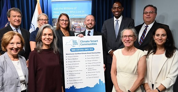 A photo of ten people from New York smiling and standing in front of a board. The board is titled "New York State Climate Smart Communities: 100 Certified Since 2014" and with a list of certified communities below. 