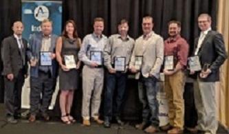 Eight men and women stand on stage holding award plaques