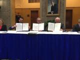 Acting Administrator Wheeler, Agriculture Secretary Perdue, and FDA Commissioner Gottleib sit at a table holding up the the papers they just signed