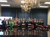 Twenty-one women and men stand behind a large conference table