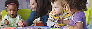 Picture of three toddlers sitting with a woman at a table playing with crayons.