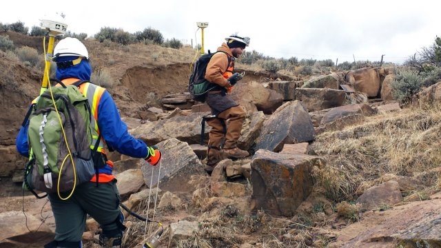 Workers carrying packs, gps locators, heavy safety gear walking over boulders.