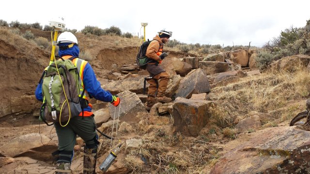 Workers walking over boulders wearing hardhats and carrying backpacks with GPS locators sticking out above their heads.