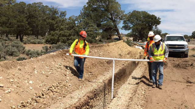 Workers carrying PVC pipe in a 'T' shape with the lower arm centered deep into an open trench on the side of a road.