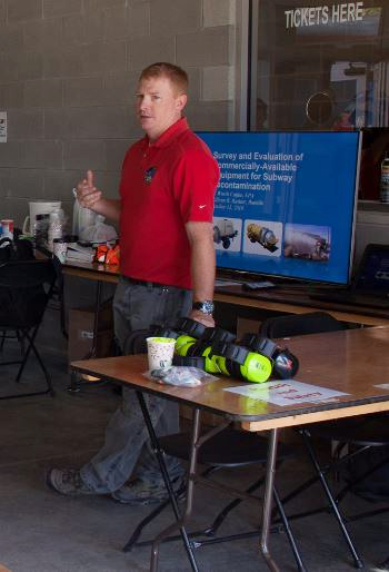 Worth stands at a table demonstrating and pointing at a poster