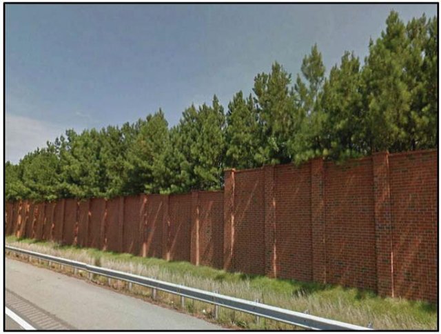 Photo shows a large brick wall with trees and vegetation extending over the top. The wall and vegetation is lining the side of a highway.