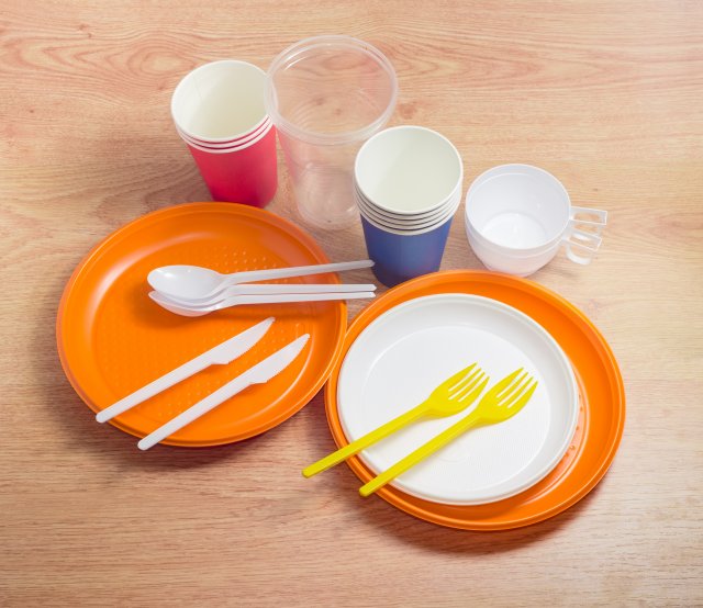 This is a picture of some nondurable goods on a wooden table, include white, plastic spoons and knives; yellow, plastic forks; white and orange paper and plastic plates; and plastic cups that are clear, white, red and blue.
