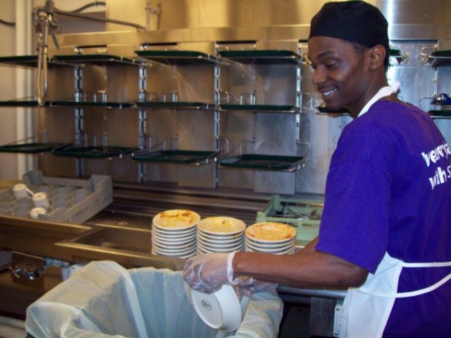 This is a picture of a gentleman cleaning dirty dishes in a industrial-type of kitchen. He's smiling as he empties contents of a bowl into a large trash bin. Three stacks of dirty dishes are on the counter behind him.