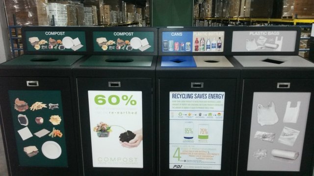 This is a picture of four different compost and recycling bins lined in a row, adjacent to each other. Each are labeled with words and pictures. From left to right, there are two compost bins, a bin for cans and plastic bottles, and one for plastic bags.