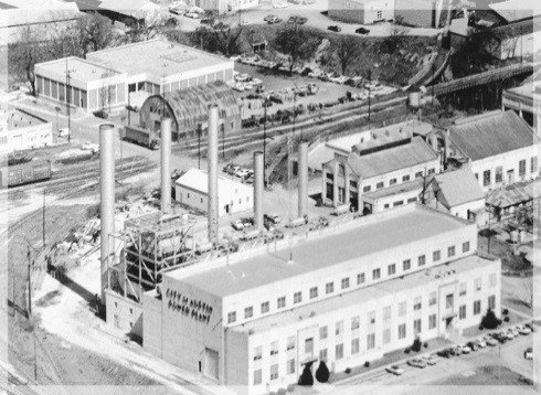 This is a black and white photo of the Seaholm Power Plant. It's an aerial shot of a factory with smokestacks. Other buildings are nearby and there is a street splitting the factory from some of the other buildings.