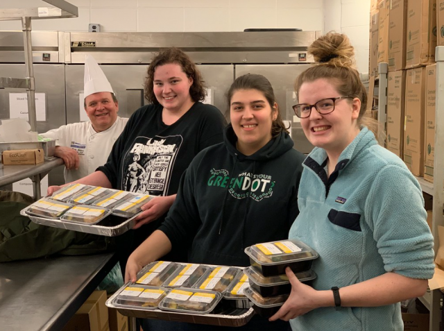 This is a photo of three UMASS Dartmouth students posing with a university chef in a facility kitchen. The students are holding food.