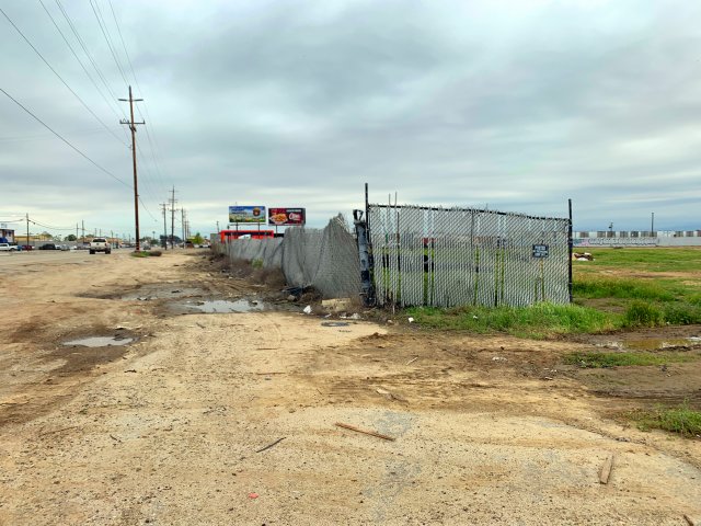 empty lot alongside a road with puddles, chain-link fence and overgrown weeds