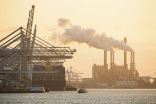 A photograph of gantry cranes and a ship with air emissions from smoke stacks in the background.