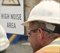 A photograph of a man wearing a hard hat with ear protection with a sign saying "High Noise Area" in the background.