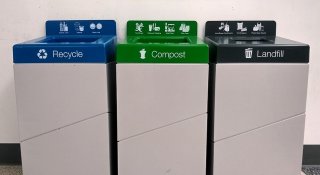Three interior airport rectangular metal bins: recycling with a blue top, composting with a green top, and landfill with a black top. Bins next to each other with that have signs with graphics showing what goes in each bin.