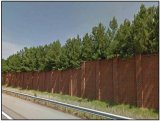 Photo shows a large brick wall with trees and vegetation extending over the top. The wall and vegetation is lining the side of a highway.