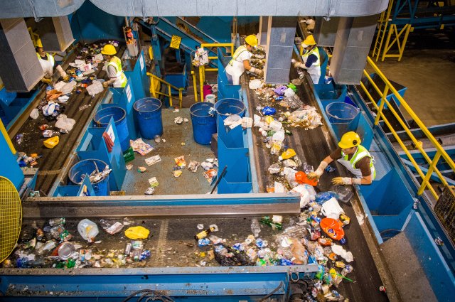 This is a photo of workers at a recycling center sorting materials that can and can't be recycled as they pass by on a conveyor belt.