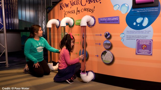Two elementary aged children engage with an indoor museum exhibit about water, featuring large transparent tubs of liquid.