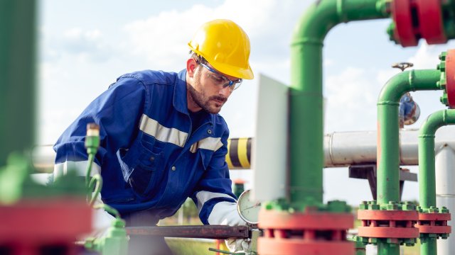 A worker dressed in blue with a yellow hard hat adjusts an outdoor pressured valve.