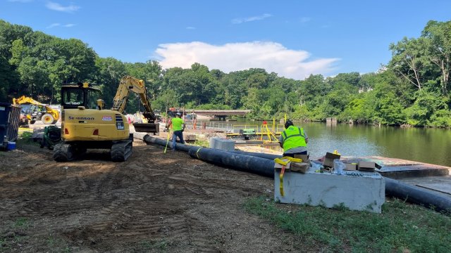 On the right, two construction workers are standing over a large pipe next to the river. A large piece of construction equipment is on the left. 
