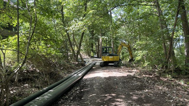 A pipeline is shown along the left side of a trail, with a worker operating a piece of earthmoving equipment with a bucket along the right side of the trail in the background. 