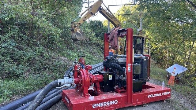 Red pump in the middle of a trail hooked up to a pipeline. Construction equipment is behind the pump on the trail.