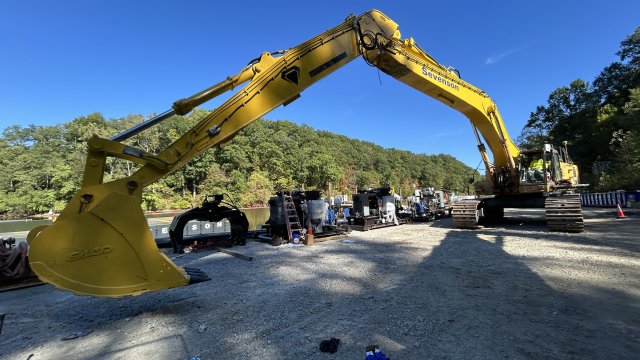 Large construction equipment with long neck is on the ground in front of river in background.