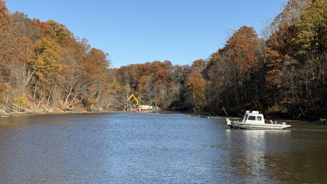 A small white boat is on a river with larger construction equipment on the water in the background. The river is surrounded by trees.