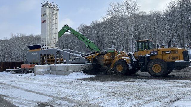 Yellow dump truck in front of a green excavator arm and white storage tower with snow on the ground.  