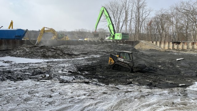 A large green excavator, a large yellow excavator, and a yellow earth mover are at work in a field of dark earth with a dusting of snow on top. The field is surrounded by brown corrugated sheet pile walls, and bare trees are in the background.