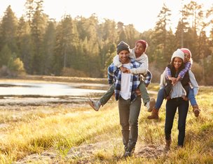 Two parents each carrying one child piggyback style through a grassy field in autumn.