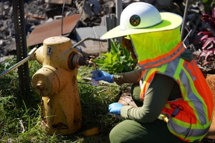 An EPA worker is seen in safety gear taking a water sample from a fire hydrant. The fire hydrant has a spigot on it to allow a small stream of water to come out