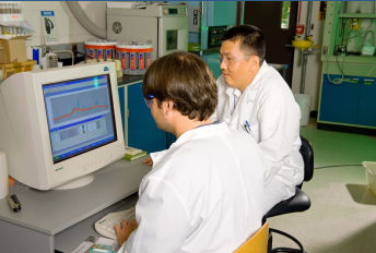 two men in white lab coats sitting in a laboratory in front of a monitor with a graph on it