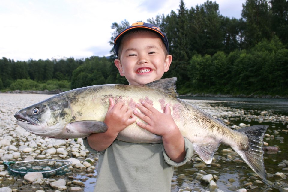 A small child smiles while holding a fresh-caught, very large salmon in his arms