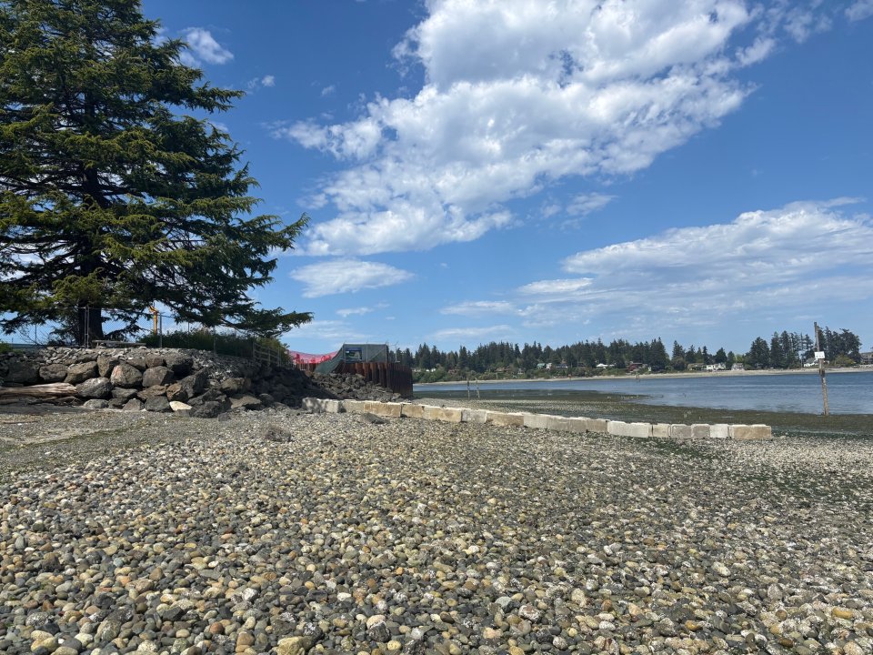 Pebbly beach in foreground with cement barrier blocking access to construction area beyond.
