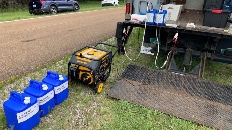 Equipment on the back of an open box truck, including blue water jugs and a generator. The equipment is used to generate bleach.
