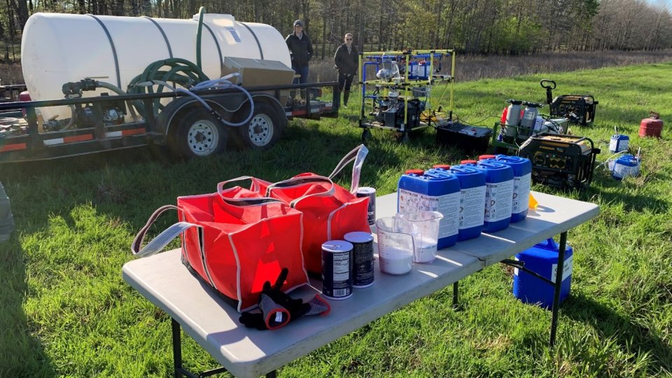 A folding table in the foreground with bleach solution being made by the WOWCart equipment. Beyond the table are the WOWCart and a trailer with a large white tank on it.