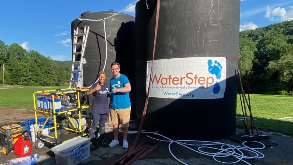 Two people give a thumbs up. They are standing in front of two large, black storage tanks labeled WaterStep. The WOW Cart is next to them.