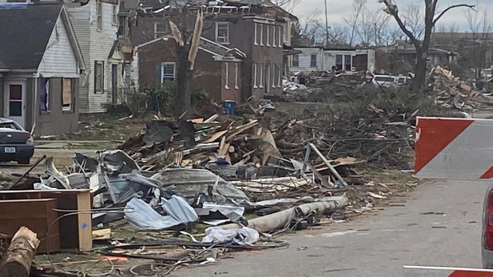 A residential neighborhood with tornado destruction. Debris and waste is piled along the road and houses in the background have tornado damage.