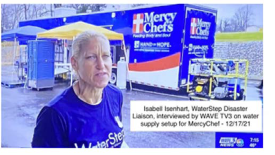 A still capture from a video, showing a WaterStep Disaster Liaison being interviewed by the news. A blue Mercy Chefs trailer is behind her.