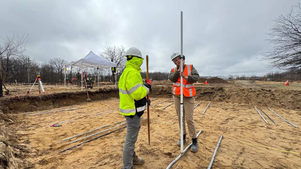 Two people in construction PPE standing outside with sections of pipe.
