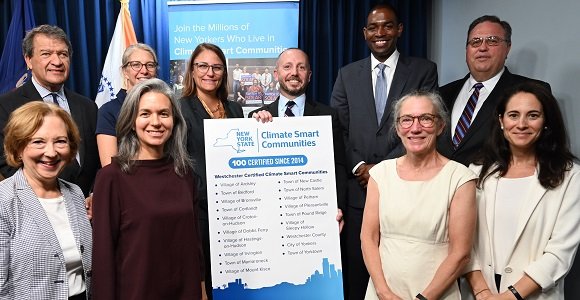 A photo of ten people from New York smiling and standing in front of a board. The board is titled "New York State Climate Smart Communities: 100 Certified Since 2014" and with a list of certified communities below.