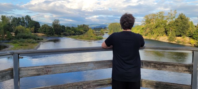 Image of a man standing on a bridge with his back to the camera. He is leaning on the railing looking at the view of the Willamette river surrounded by trees with mountains in the background