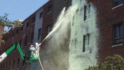 Researchers wearing white hazmat suits, standing on a ladder, spray foam on a brick building as part of a cleanup demonstration.