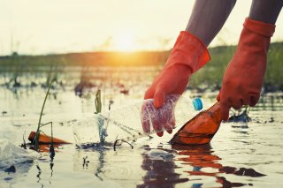 This is a picture of two orange gloved hands pulling an empty plastic water bottle and a glass bottle out of a water body surrounded by marsh. A soft yellow sun is setting in the background.