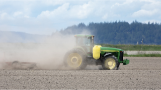 A green and yellow tractor driving across a dirt field and kicking up dust. Hill in background
