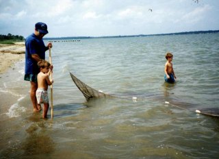A young Jake seining for fish with his father in the shallows of an estuary. His brother stands in the water up to his knees on the other side of the net. 