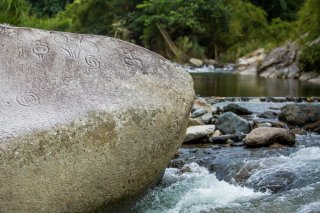 A rock with Taino carvings sits next to a stream in a forest.