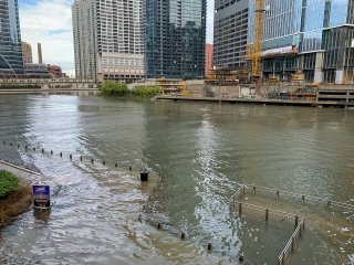 The Chicago River floods over its banks, with water covering the walking paths next to the riverbank and skyscrapers in the background.