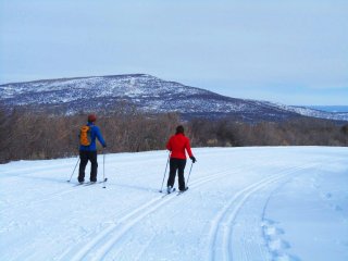 Two people cross-country skiing on snow with a small mountain in the background.